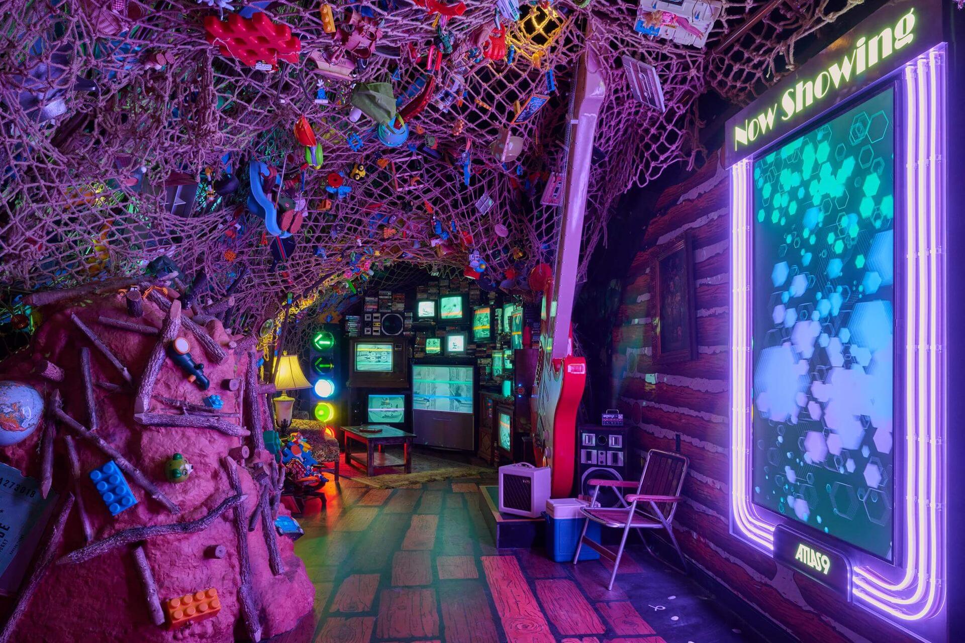 A cabin-like corridor overgrown with colorful crocheted netting, cluttered walls of screens, and a neon-framed “Now Showing” display