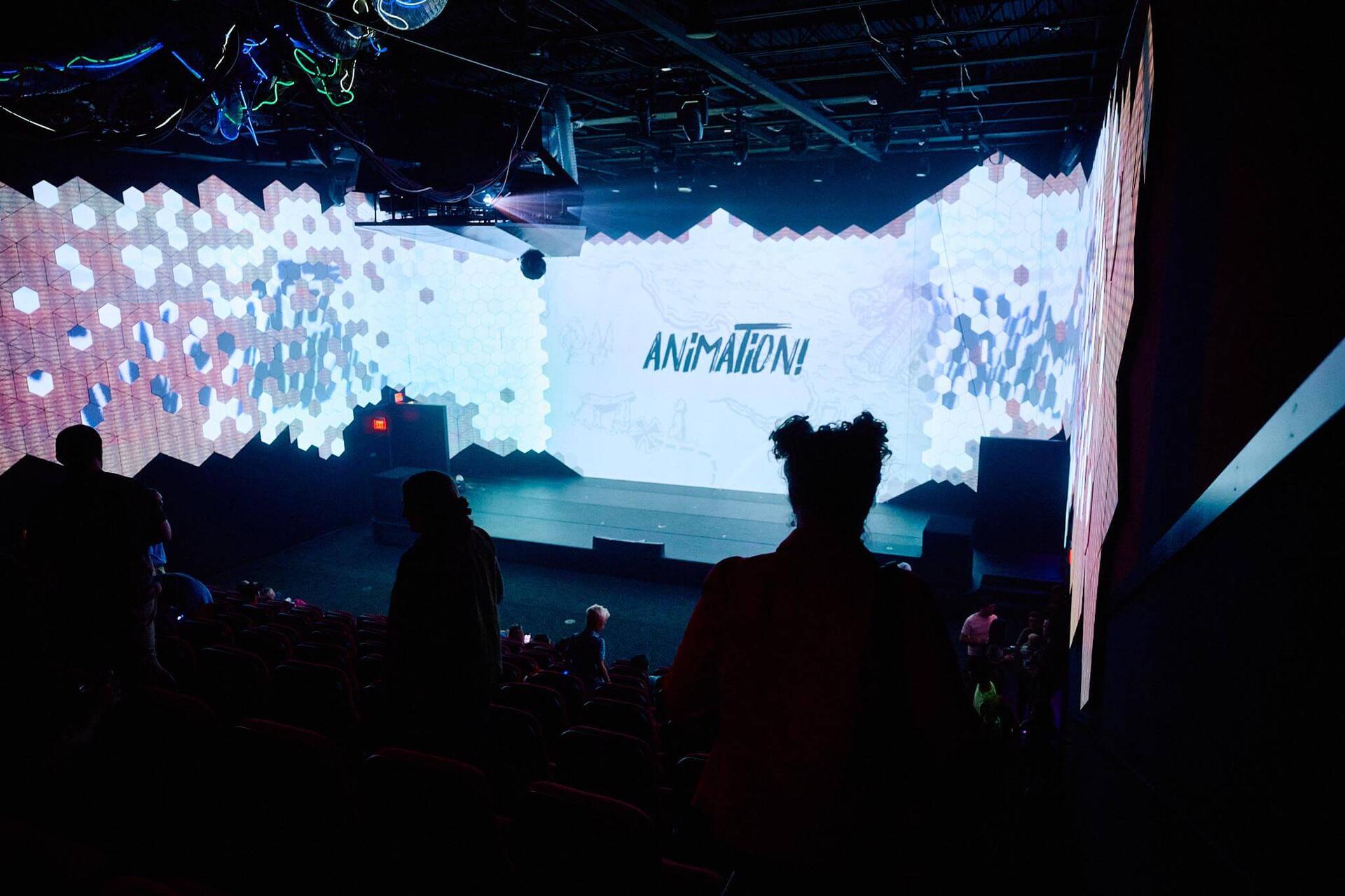 An audience watching an animated presentation on a wide paneled projection wall in the theater