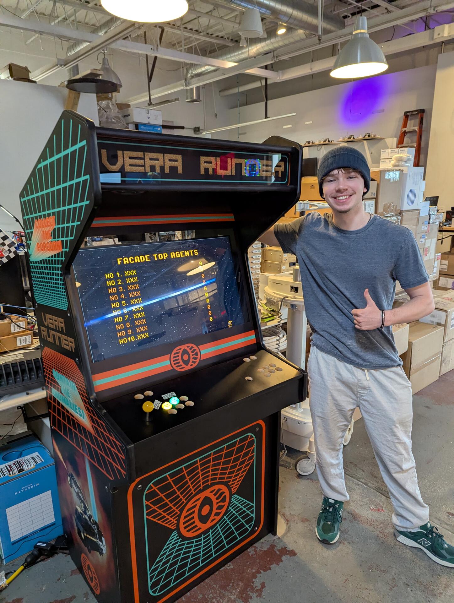 A team member giving a thumbs up next to the Vera Runner custom arcade cabinet in the workshop